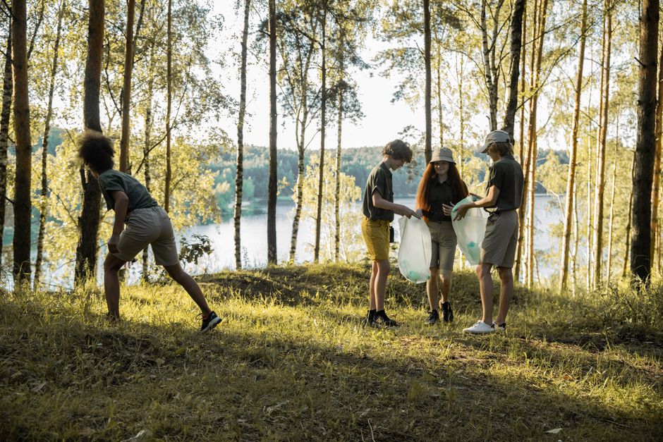 Four young individuals participating in a litter pick-up activity in a natural outdoor setting near a body of water, surrounded by tall, slender trees with yellow-green leaves and a backdrop of distan