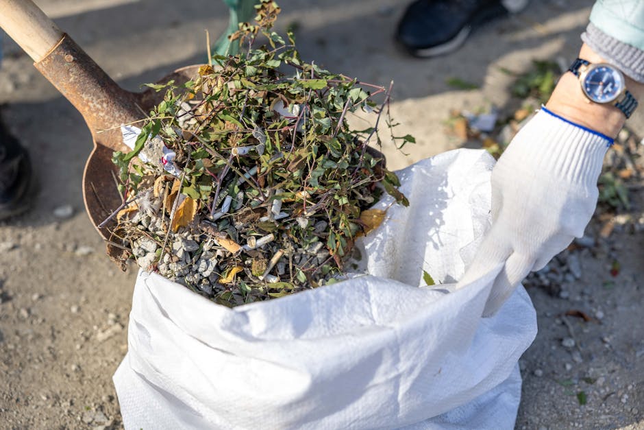 A person wearing a white glove with a blue watch on their wrist is seen holding a large white waste bag filled with garden debris, including small green plants, twigs, dried leaves, and bits of gravel